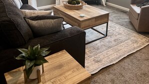 Una sala de estar con un sofá marrón oscuro, una mesa de centro de madera, una pequeña planta en maceta, un televisor sobre un mueble bajo y una ventana con cortinas blancas.