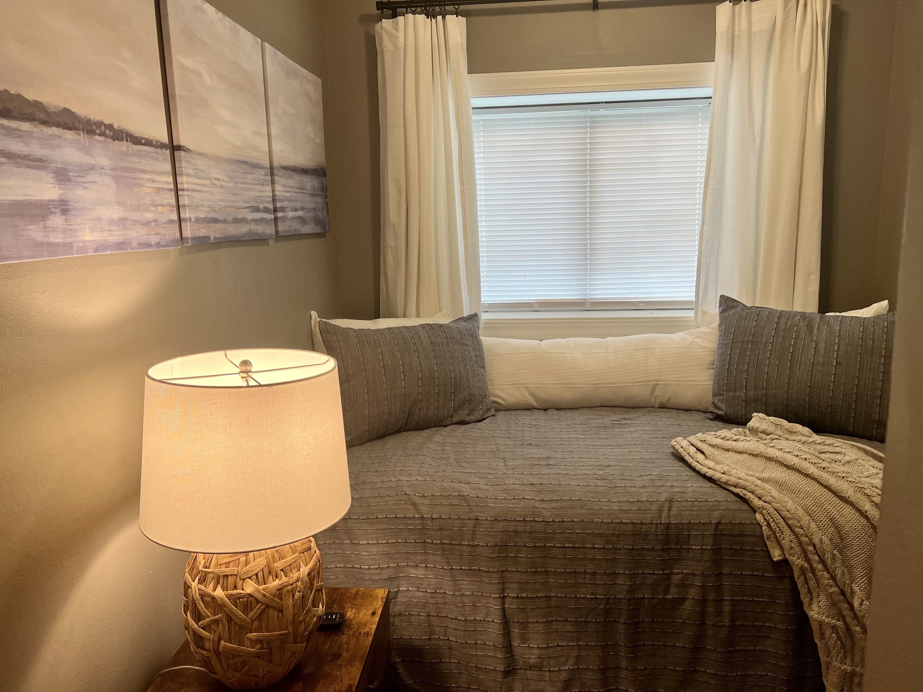 A bedroom with a bed, a lamp, a window with curtains, and a painting on the wall.