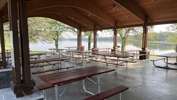Un área de picnic techada con mesas, bancos, una chimenea de piedra y vista a un lago.