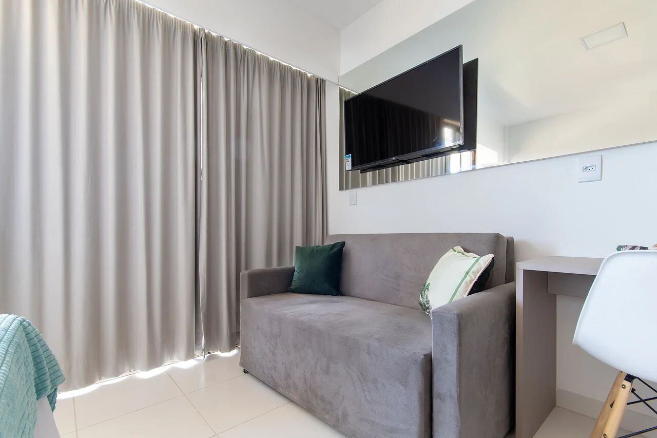 A modern living room with a grey sofa, a flat-screen TV mounted on the wall, and a large window with curtains.