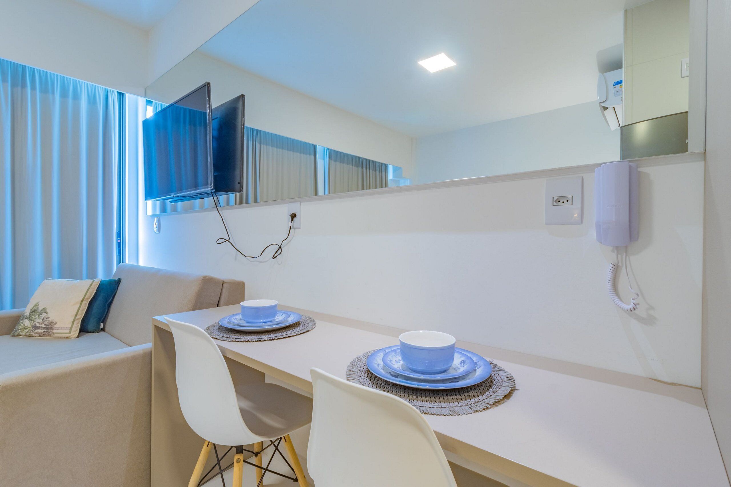 A hotel room with a dining table set for two, a sofa, a TV mounted on the wall, and a phone.