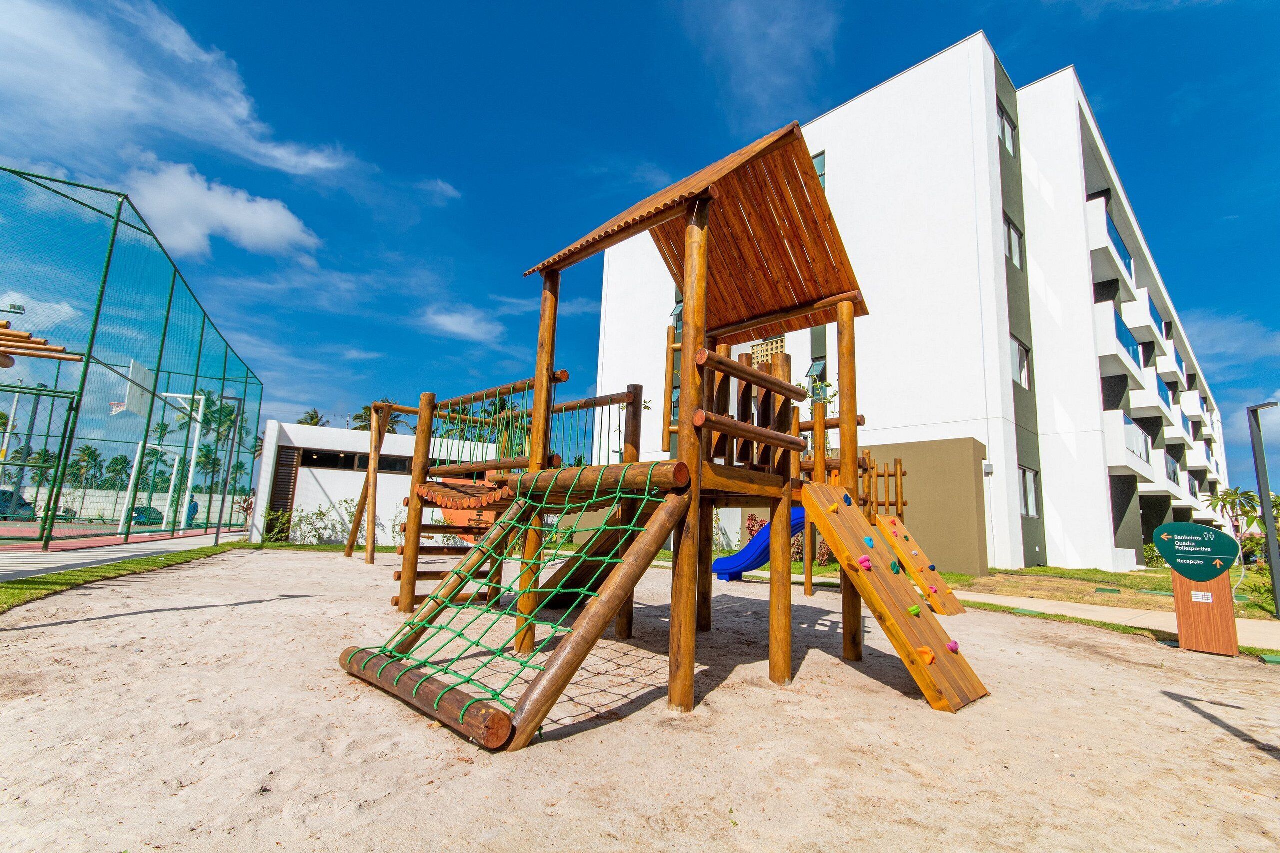 A playground with a wooden climbing structure and a slide, surrounded by a sandy area and a modern building with balconies.