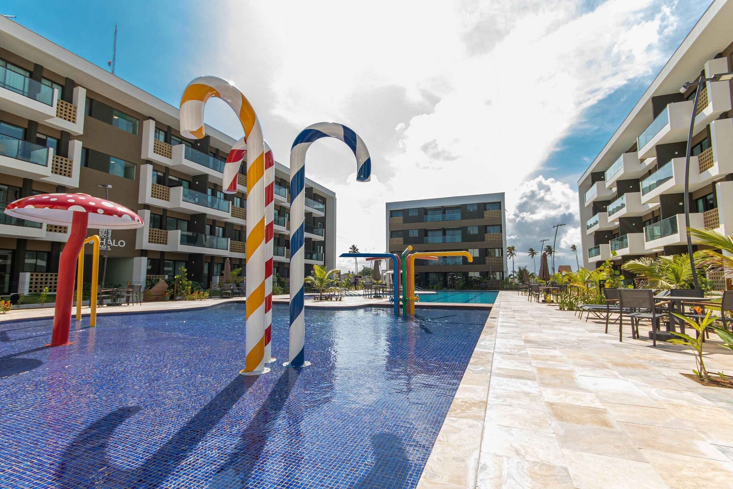 A swimming pool with colorful candy cane decorations, surrounded by modern buildings with balconies and a clear sky.