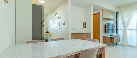Une chambre d’hôtel moderne avec une table à manger en marbre, des chaises beiges, un téléviseur et un meuble en bois.