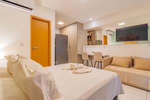 Quarto de hotel com cama, sofá, escrivaninha com cadeira, televisão de tela plana e área de cozinha compacta.
