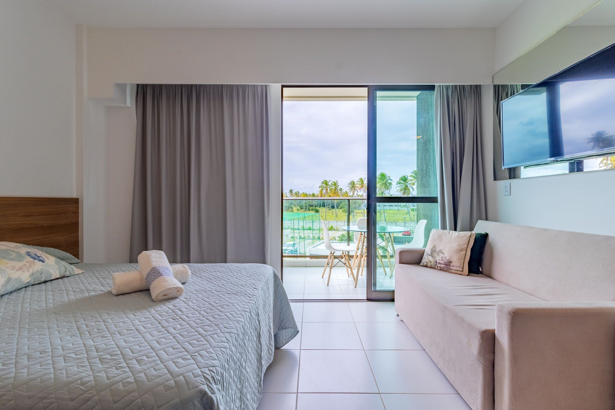 A hotel room with a bed, a sofa, a TV, and a balcony with a view of palm trees.