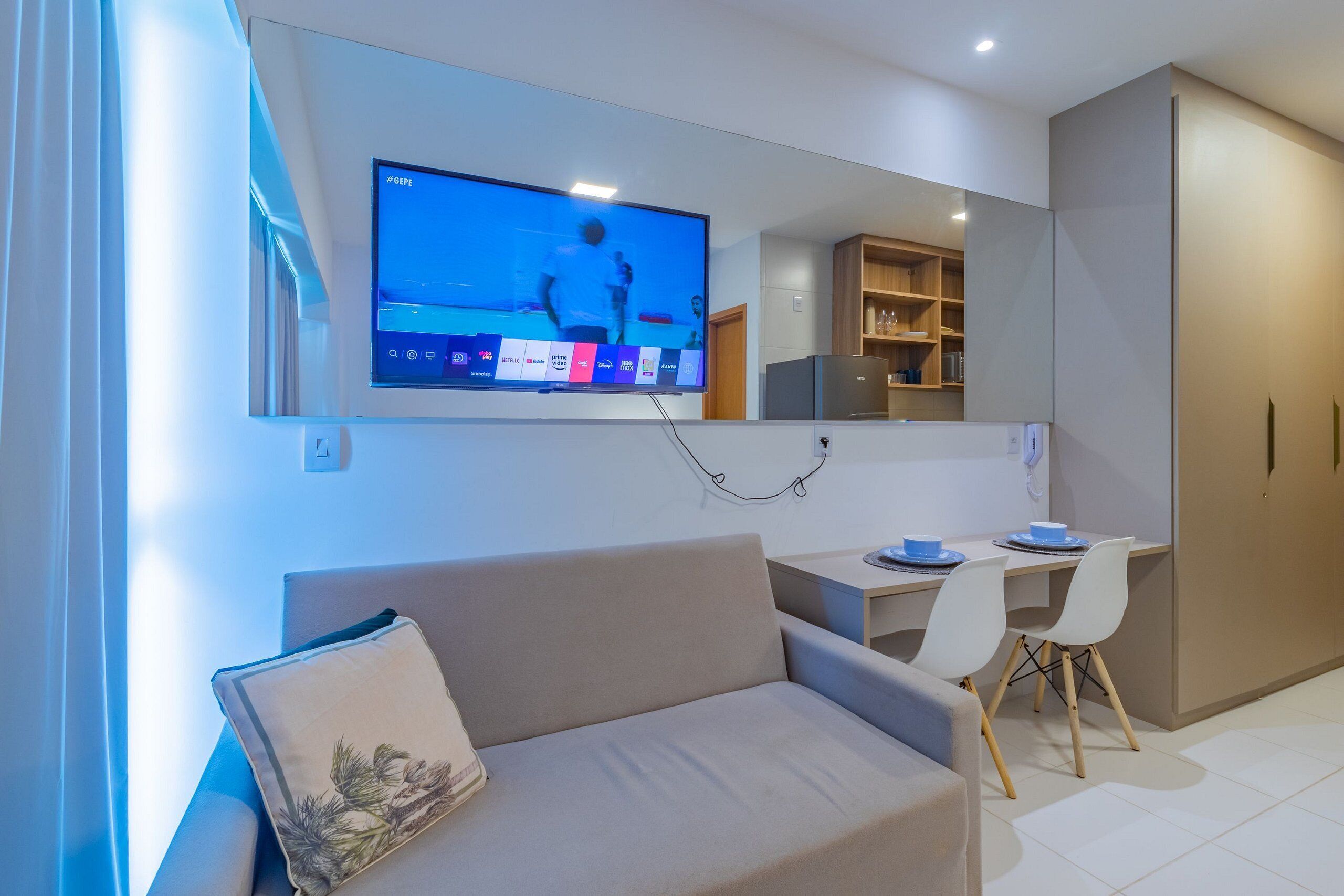Un salon moderne doté d’un téléviseur à écran plat fixé au mur, d’un canapé agrémenté d’un coussin décoratif, d’un coin repas avec une table et des chaises, et d’une cuisine équipée de placards et d’un réfrigérateur.