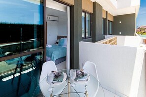 Un balcon avec une table et des chaises en verre, un lit recouvert d’une couverture bleue et une vue sur le paysage urbain.
