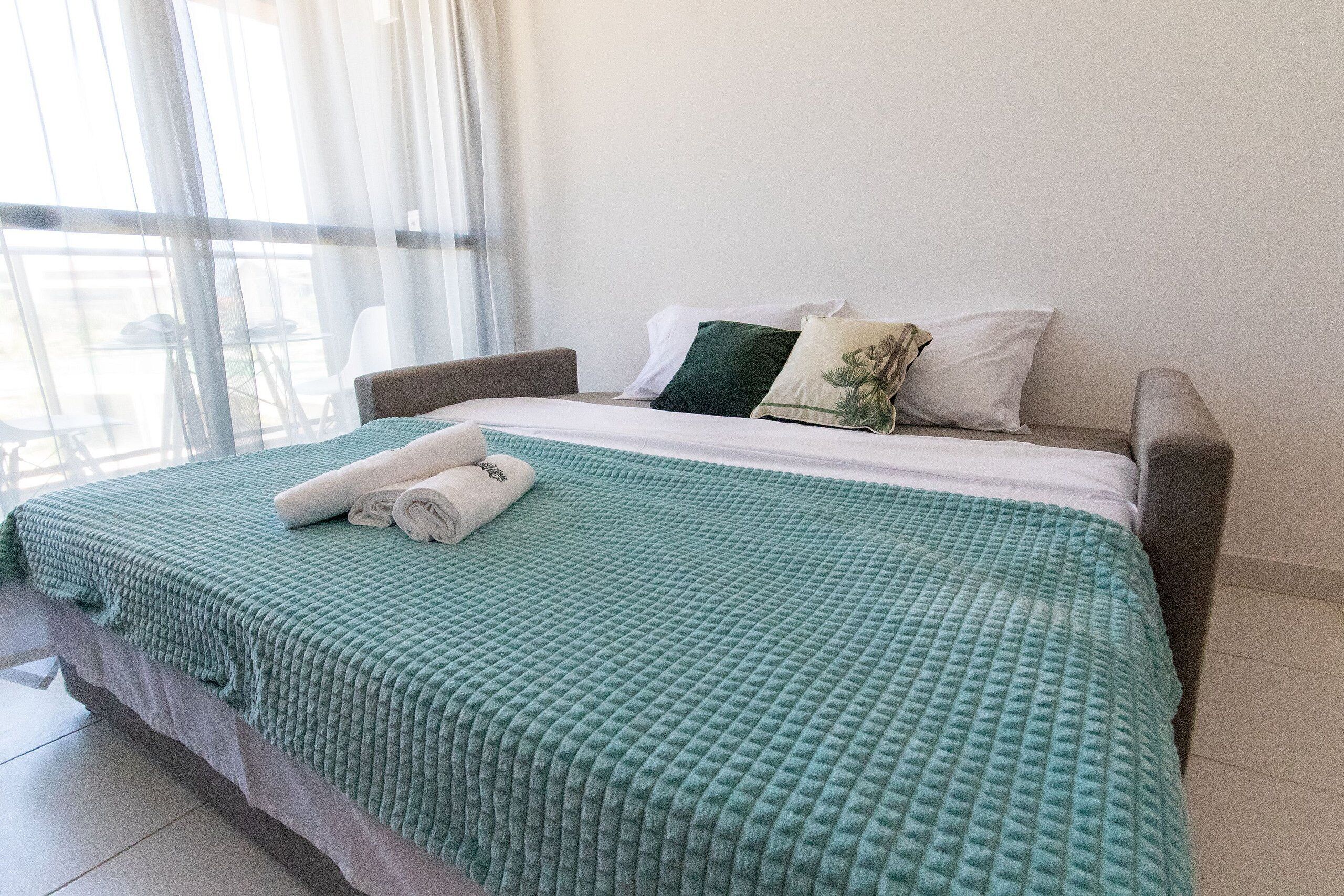 Un lit bien fait, avec une couverture verte à motifs, des oreillers blancs et une serviette enroulée.