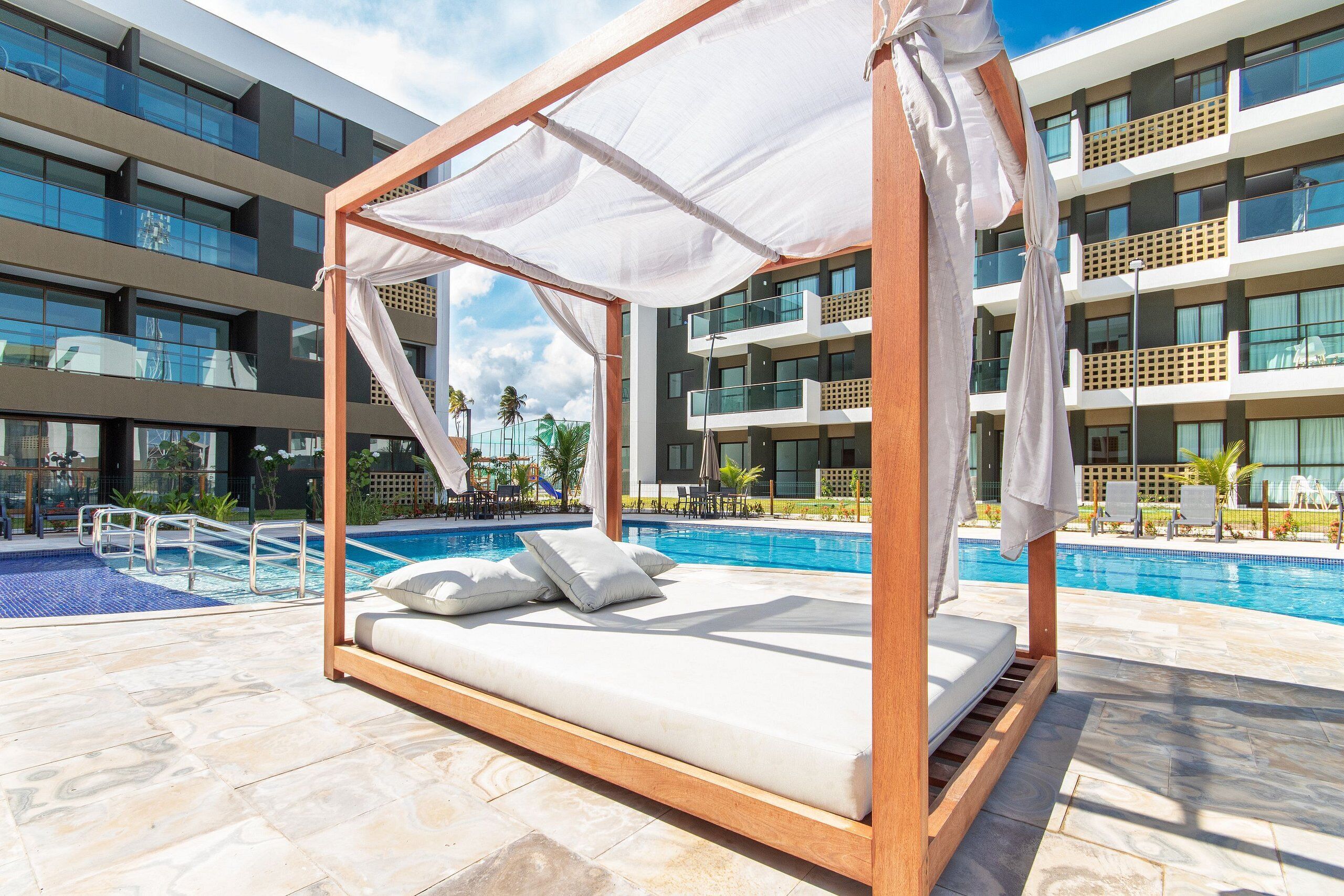 Un espace piscine privé doté d’un lit à baldaquin en bois, d’une piscine et d’un bâtiment moderne en arrière-plan.