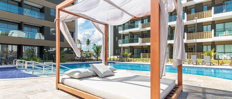Un espace piscine privé doté d’un lit à baldaquin en bois, d’une piscine et d’un bâtiment moderne en arrière-plan.