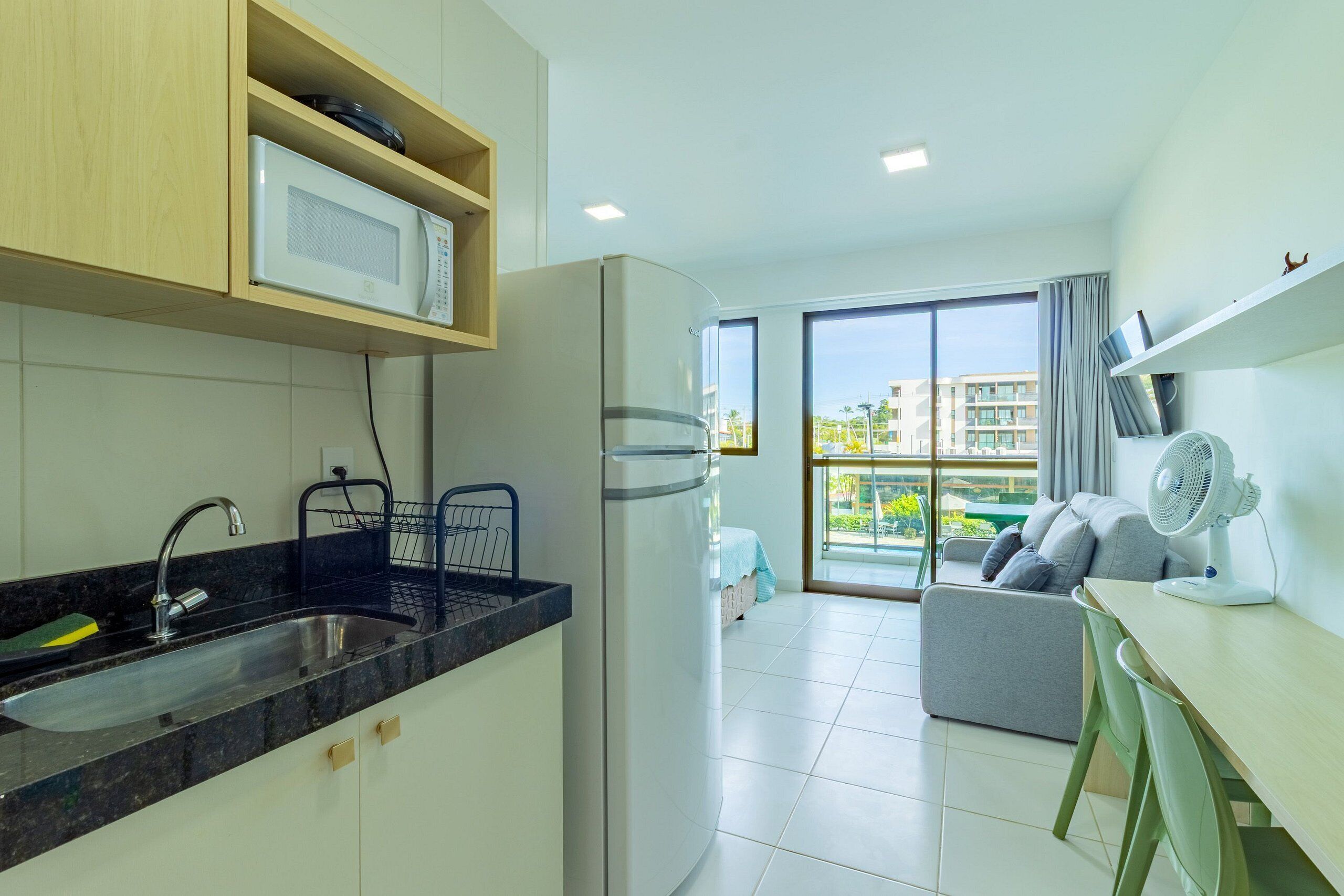 Uma cozinha moderna com micro-ondas embutido, pia e geladeira. Há uma varanda com vista para edifícios e um ventilador sobre a mesa.