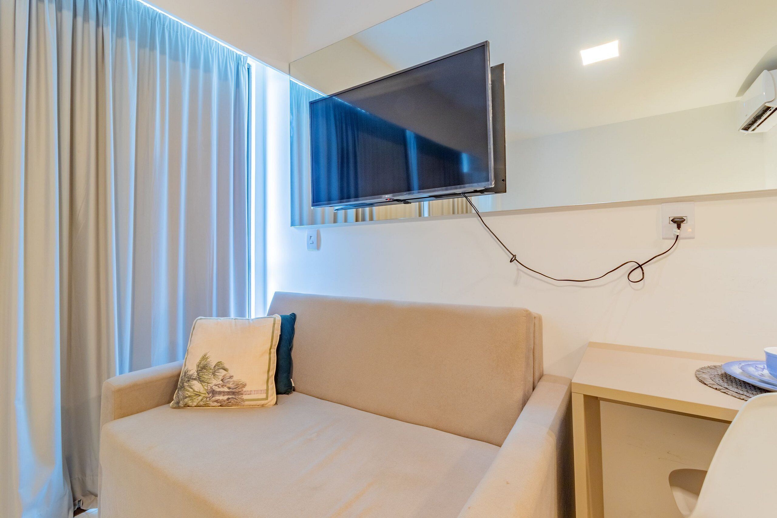 Habitación de hotel con sofá, televisión instalada en la pared, una mesita y una ventana con cortinas.