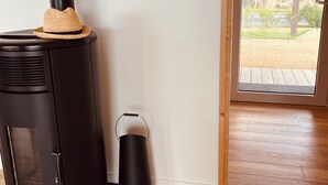 Una estufa de leña moderna y compacta con una tapa superior, un bote de basura negro, y una vista de una terraza de madera y una casa a través de una puerta de vidrio.