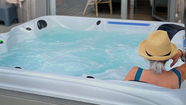Un jacuzzi con una persona adentro, una terraza de madera y una puerta de cristal que da a un espacio exterior con una silla y una mesa.