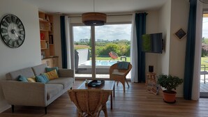 Una sala de estar moderna con un sofá, sillas de mimbre, una mesa de centro, una estantería, un televisor y vistas a un jacuzzi y un jardín.