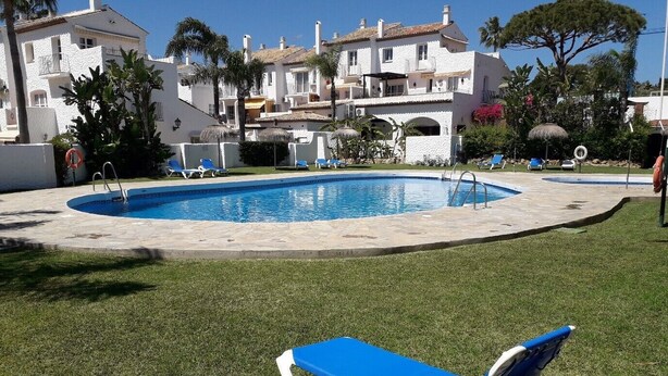 A swimming pool with lounge chairs, surrounded by a well-maintained lawn and a white building with balconies.
