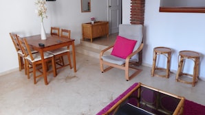 A dining area with a wooden table and chairs, a cushioned chair with a pink pillow, a small wooden cabinet, and a glass-top table with a purple rug.