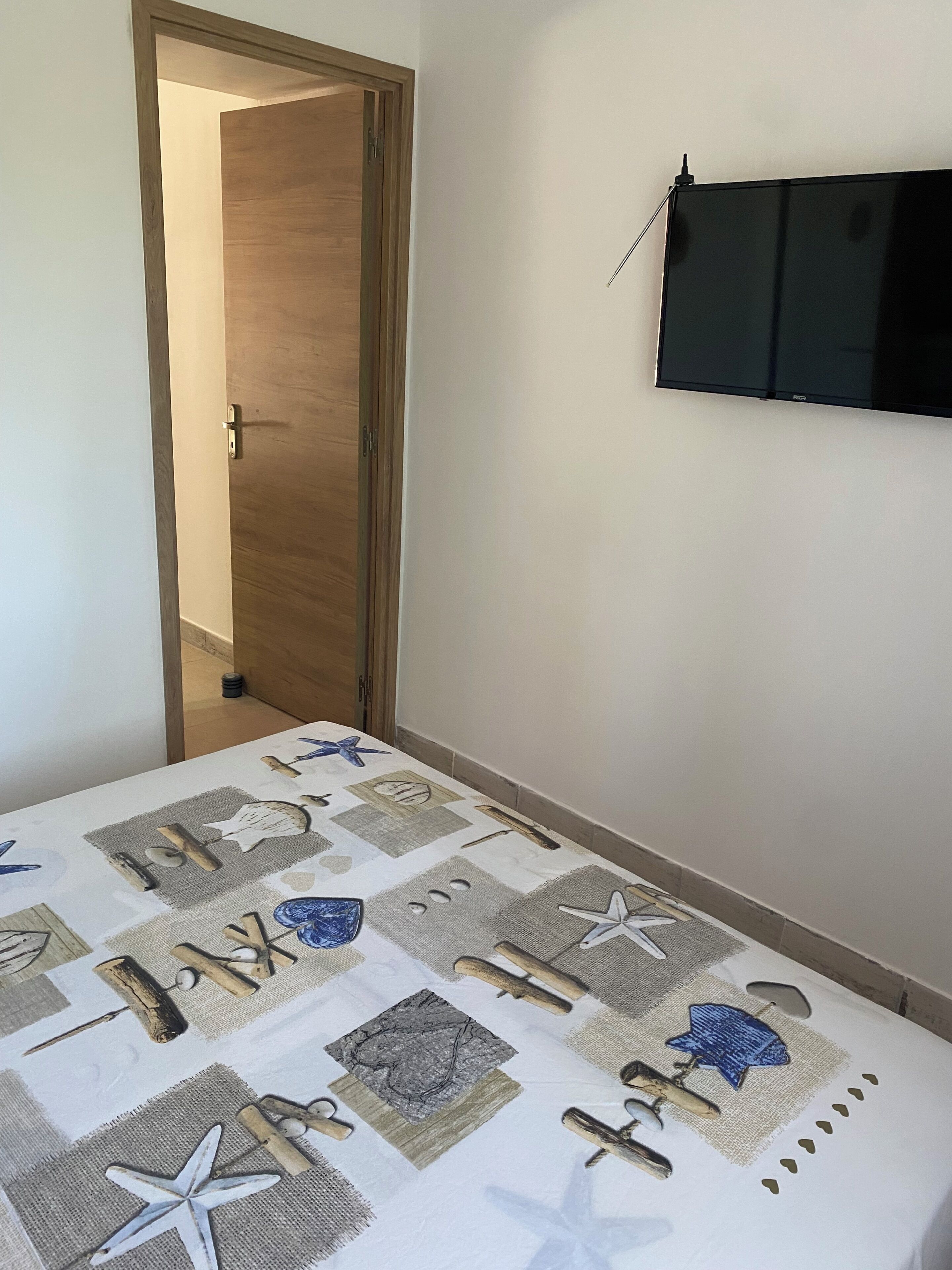 Une chambre avec un lit, une porte en bois, un téléviseur fixé au mur et une table recouverte d’une nappe sur le thème de la marine.