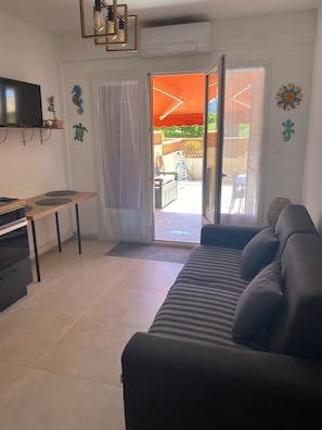 A living room with a sofa, a TV, a small table, and a sliding glass door leading to an outdoor patio. - Lava Plage - Feet in the water (appietto)