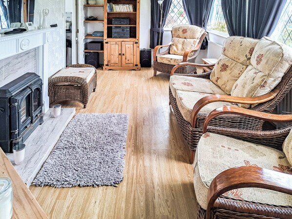 A cozy living room with a fireplace, wicker furniture, a bookshelf, and a window with curtains.