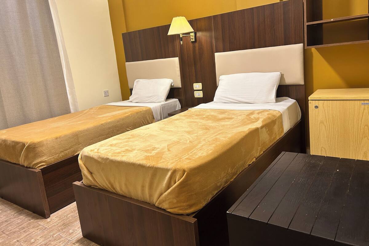 A hotel room with two single beds, a wooden nightstand, a small table, and a wooden wardrobe.