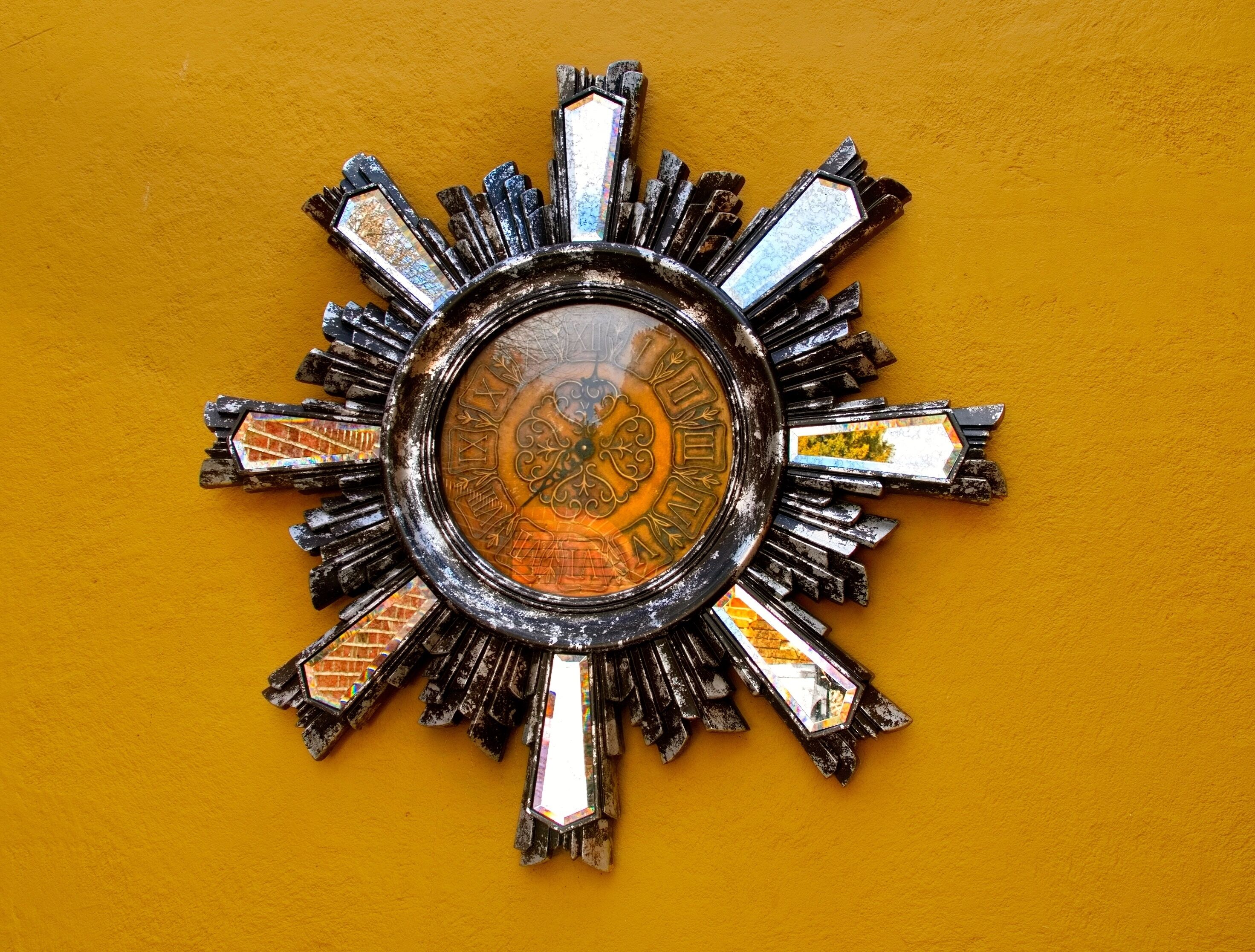 A sun-shaped clock with a central dial and multiple reflective surfaces on a yellow wall.