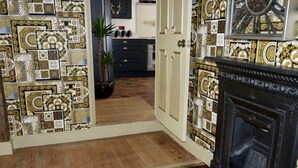 Una habitación con paredes de intrincado diseño dorado y blanco, suelo de madera, chimenea negra y una puerta que da a otra estancia.