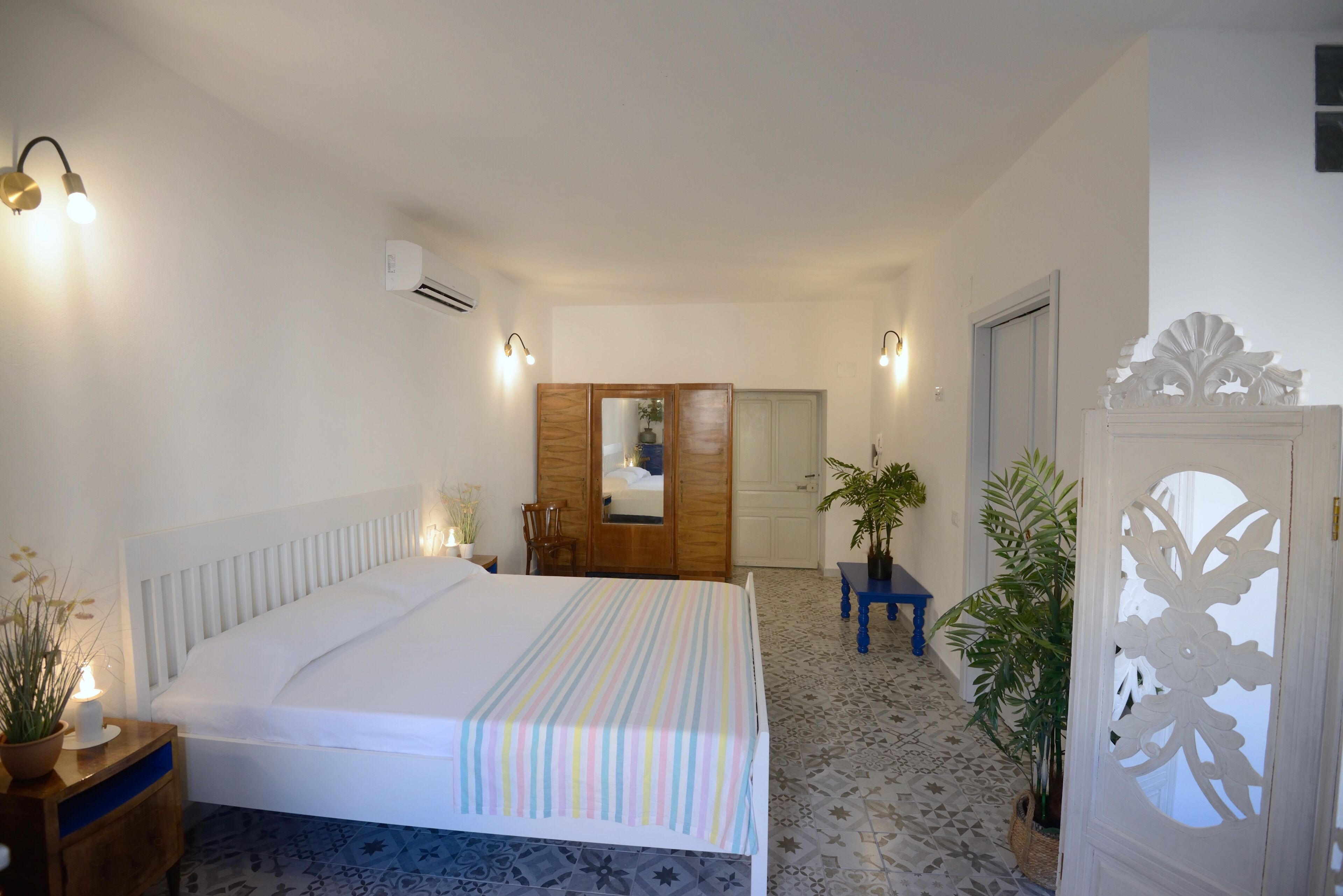 Quarto com cama, mesa de cabeceira, luminária, aparelho de ar condicionado e porta com detalhe decorativo.