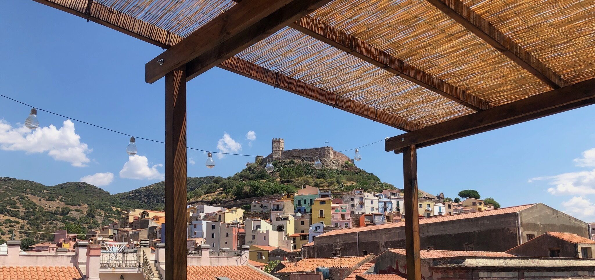 Blick auf eine Küstenstadt mit bunten Gebäuden, einer markanten Struktur auf einem Hügel und einer Holzpergola mit Lichterketten.