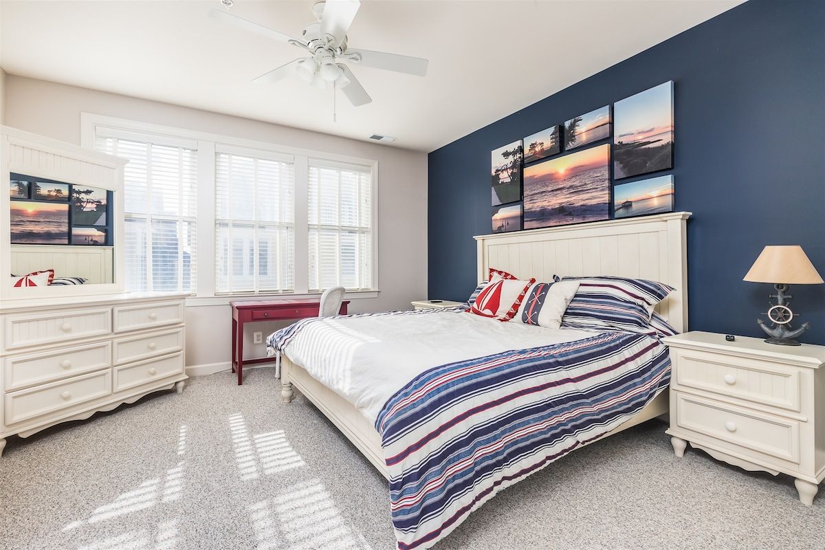 Une chambre à coucher comprenant un lit, un bureau, une chaise, une commode, une table de chevet et un ventilateur de plafond.