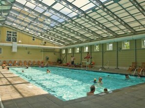 An indoor swimming pool with a glass ceiling, surrounded by lounge chairs and a tiled floor.
