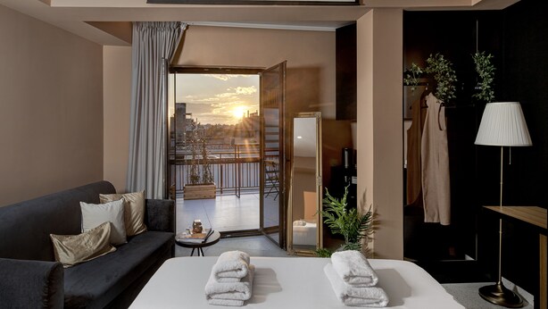 A hotel room with a bed, a sofa, a table with towels, a lamp, and a view of the city at sunset. - Clyde Athens by Nin&Bau (Athens)