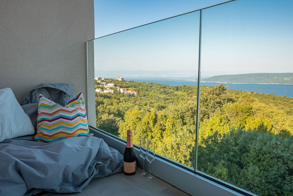 A balcony with a bed, a bottle of champagne, and two glasses. The balcony overlooks a scenic view of a coastal area with greenery and buildings in the distance.