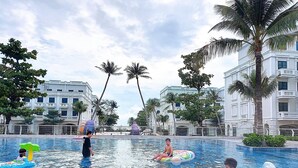 A swimming pool area with multiple people, some on inflatable rings, surrounded by palm trees and modern buildings. - Kalia Hotel (Phu Quoc)