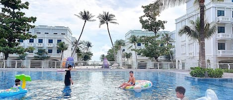 A swimming pool area with multiple people, some on inflatable rings, surrounded by palm trees and modern buildings.