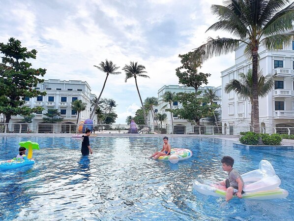 A swimming pool area with multiple people, some on inflatable rings, surrounded by palm trees and modern buildings.
