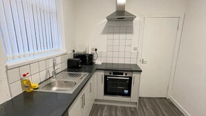 Cozinha compacta com bancada preta, armários brancos, forno embutido e janela com persianas.