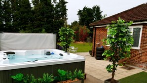A hot tub with two planters on either side, a brick house in the background, and a lawn. - Deluxe 4/5 Bed Hot Tub Cinema Room 15 (Halstead)