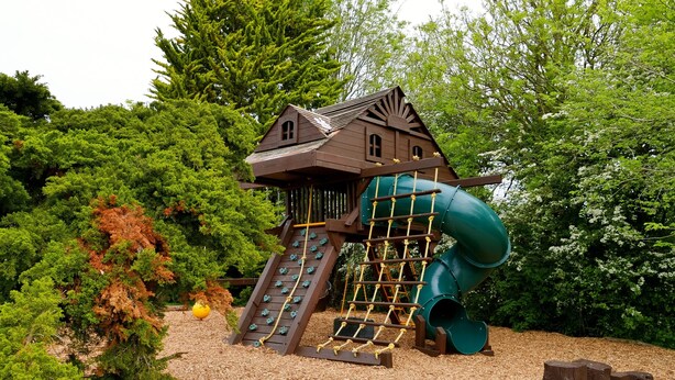 A wooden playhouse with a slide and climbing net, surrounded by trees and mulch. - Deluxe 4/5 Bed Hot Tub Cinema Room 15 (Halstead)