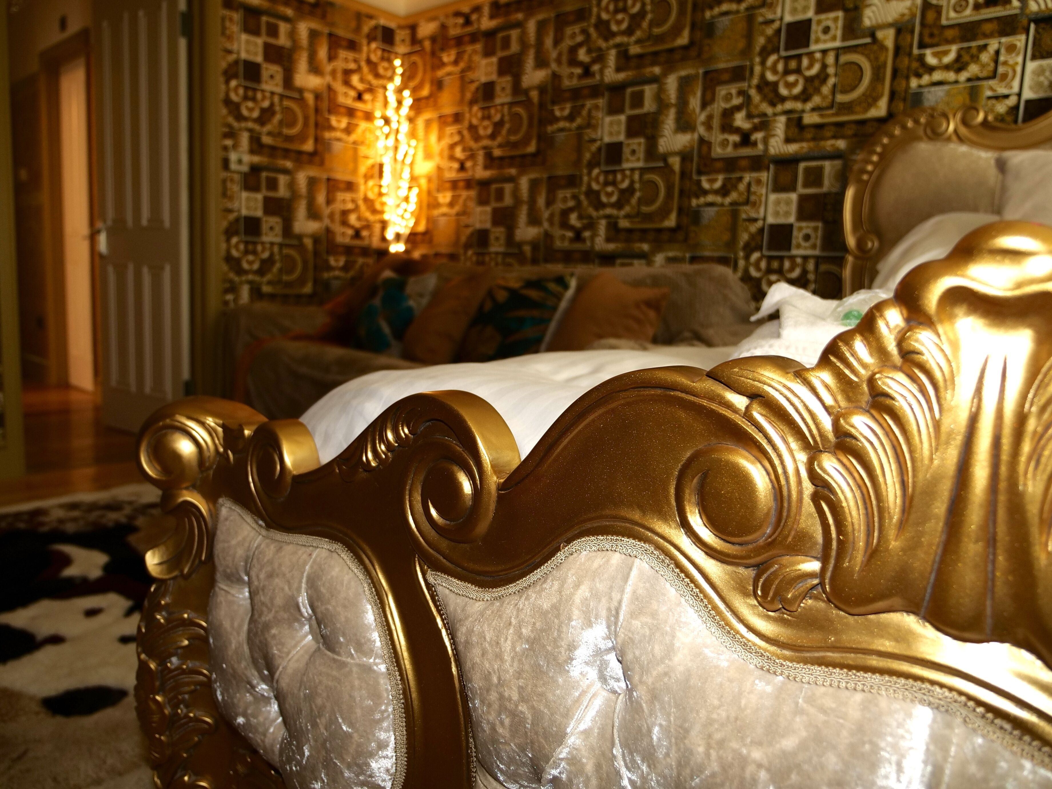 A luxurious bed with a gold headboard and white bedding, set against a patterned wallpaper and a chandelier.