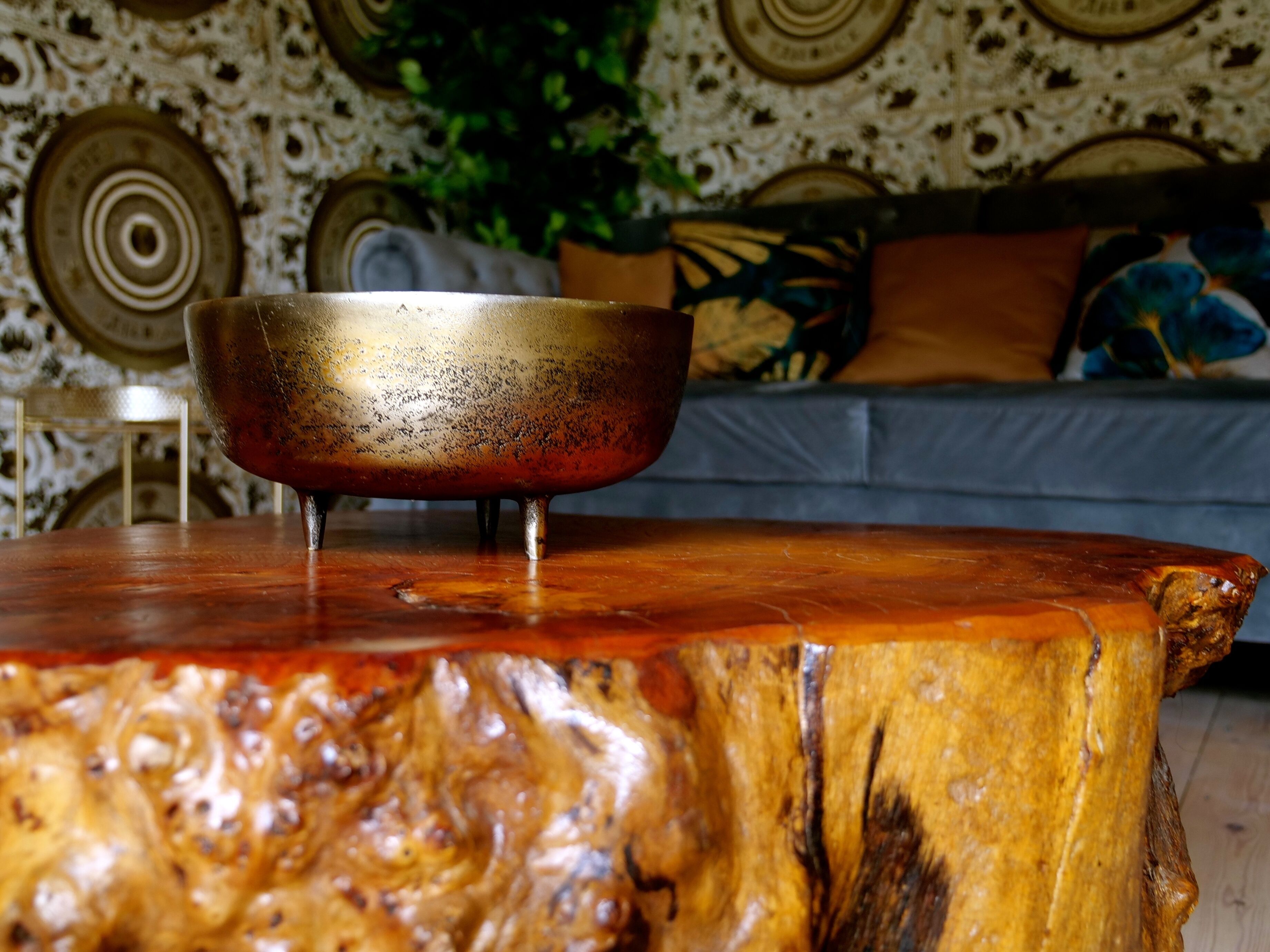 A wooden table with a brass bowl on it, set against a patterned wall and a sofa with cushions.