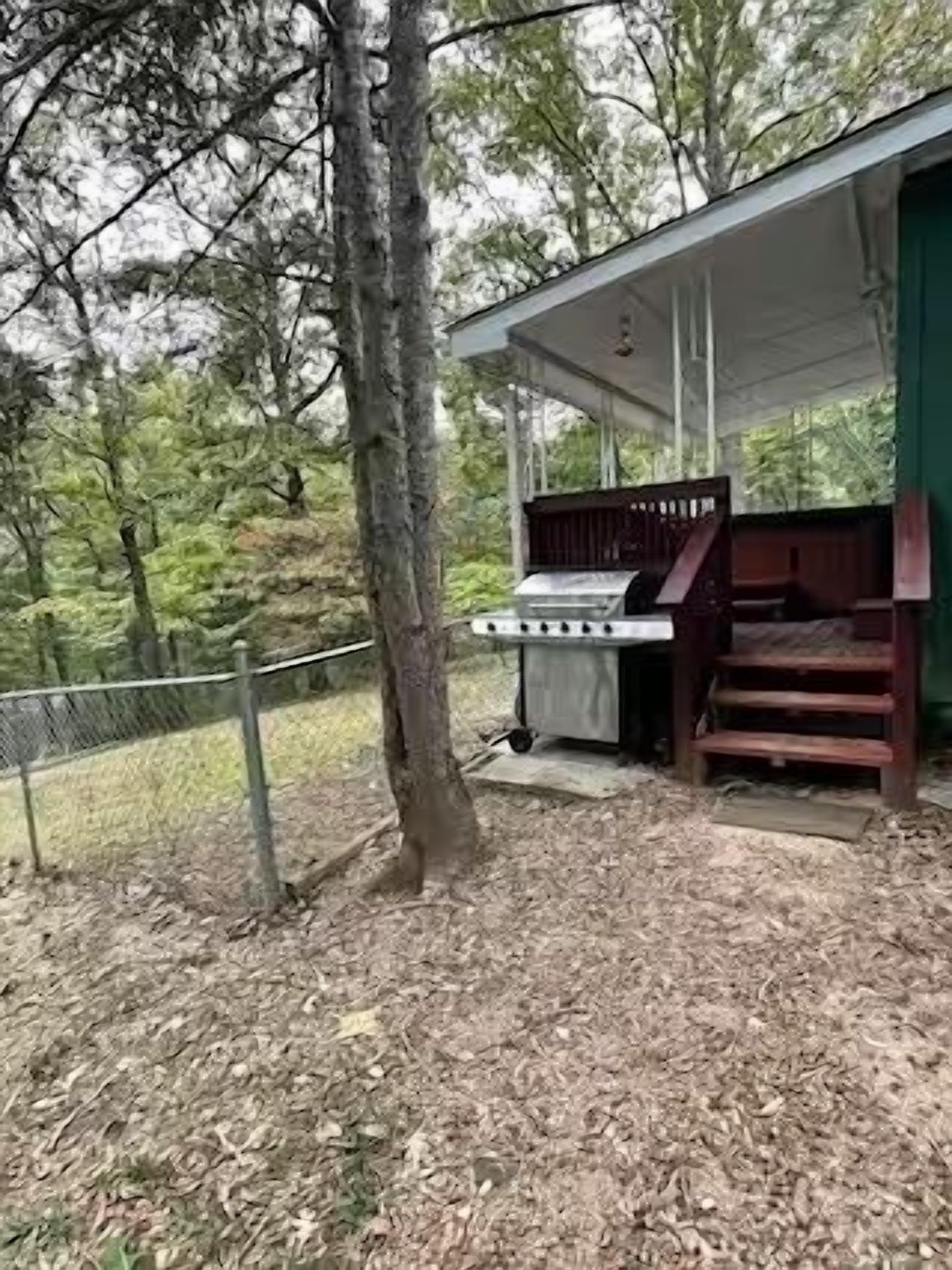 A covered outdoor grill area with a wooden bench, surrounded by trees and a fence.
