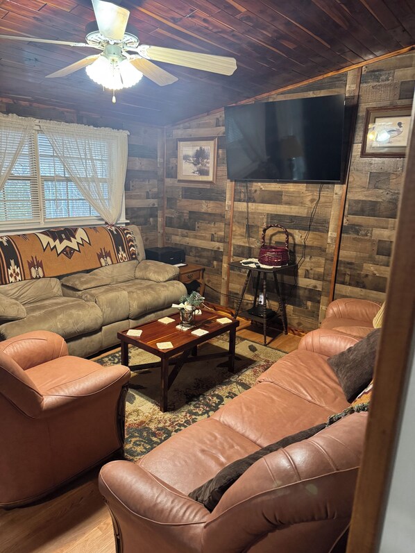 A cozy living room with a wooden ceiling, a ceiling fan, a large TV, a sofa, a coffee table, and a patterned rug.