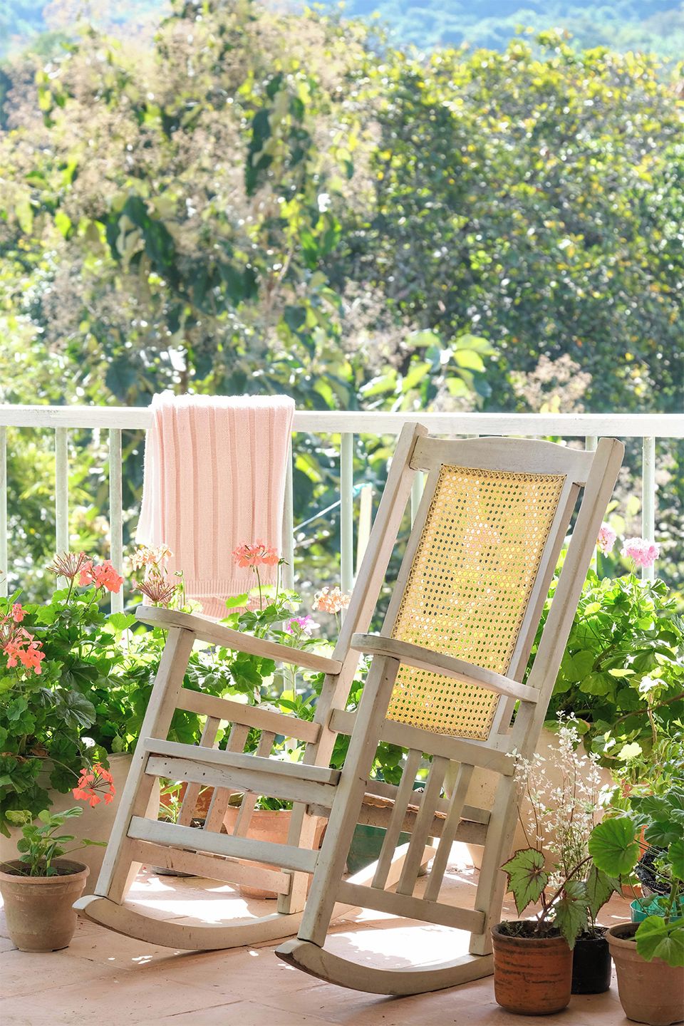 A wooden rocking chair with a woven backrest on a balcony with potted plants and a pink towel hanging on a railing.