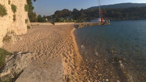 Une plage bordée d’un muret de pierres, avec une eau cristalline et un voilier à l’horizon.