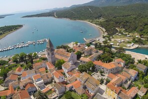 Vue aérienne d’une ville côtière, avec un clocher d’église imposant, un port de plaisance rempli de bateaux et des immeubles résidentiels aux toits recouverts de tuiles rouges.