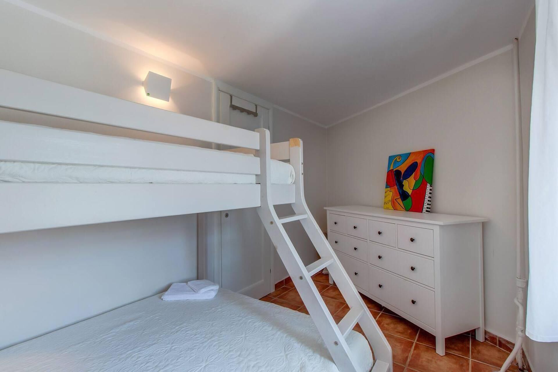 Una cama litera con escalera, un aparador blanco y un cuadro colorido en la pared.