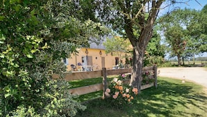 A garden area with a wooden fence, a tree with pink flowers, and a building with outdoor seating. - Bungalow on a horse farm (Yèvres)