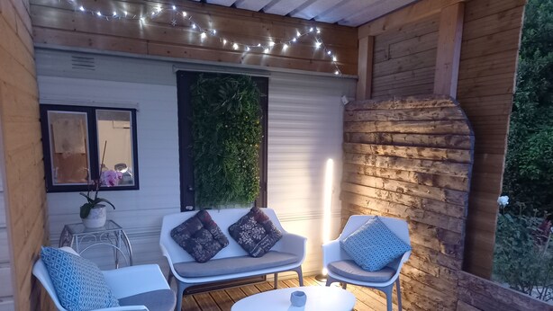 A wooden deck with a seating area featuring two chairs and a small table, a decorative wall with greenery, and string lights. - Bungalow on a horse farm (Yèvres)